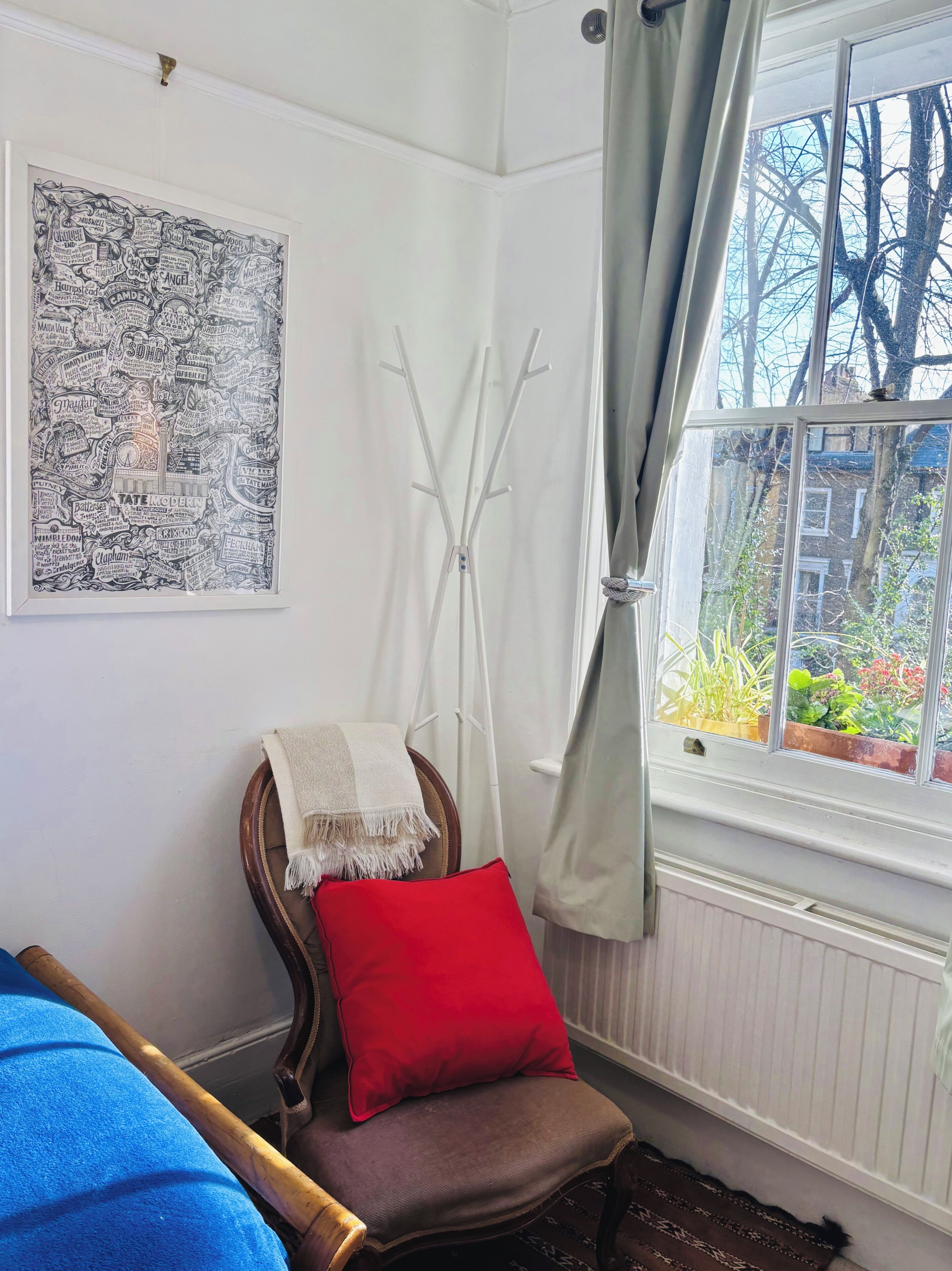 A room with a chair, a red pillow, a coat rack, a window with a view of trees and buildings, and a framed map on the wall.