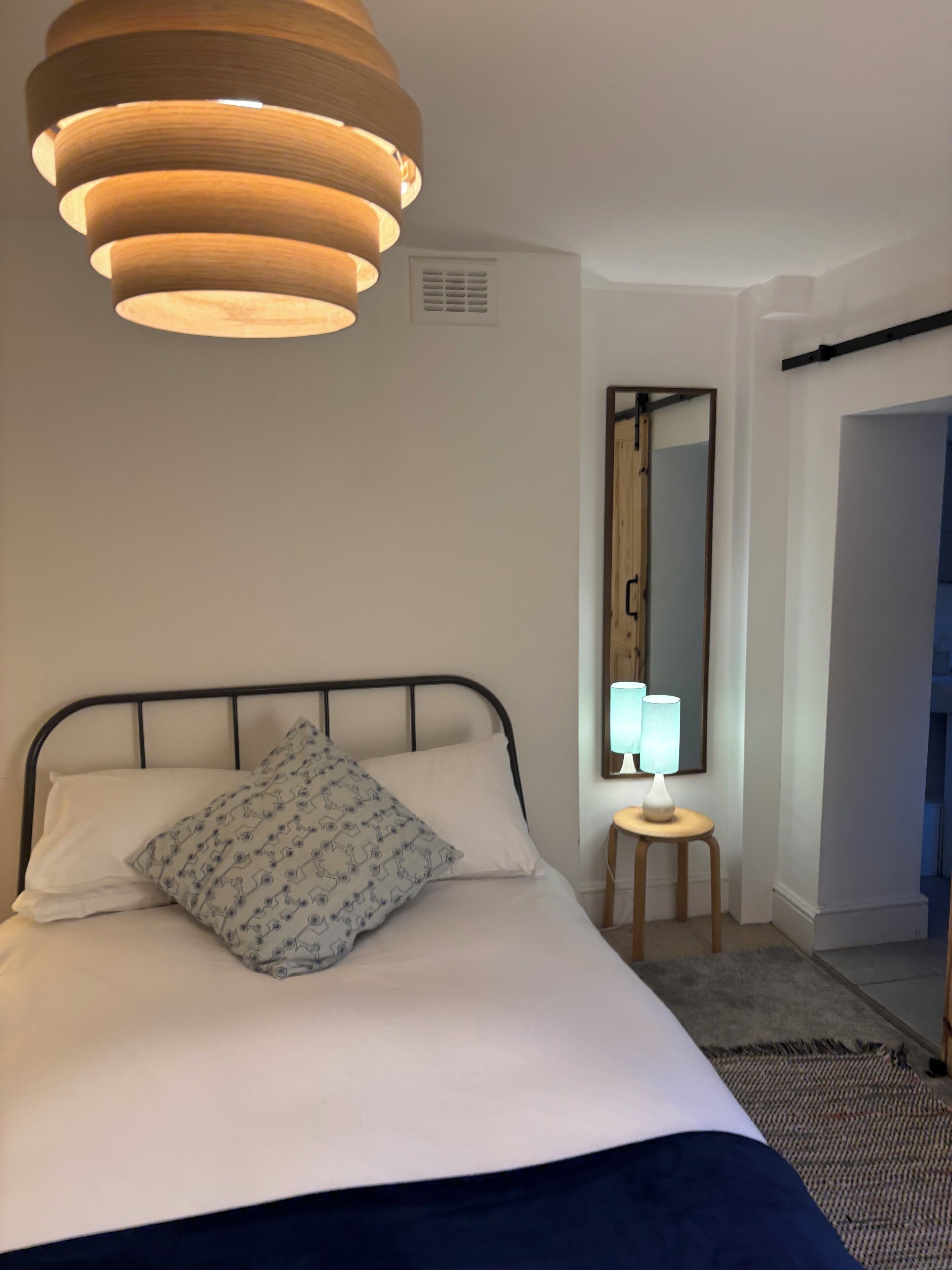 Una cama bien hecha con un almohadón estampado, una lámpara de noche, un espejo y un aplique colgante.