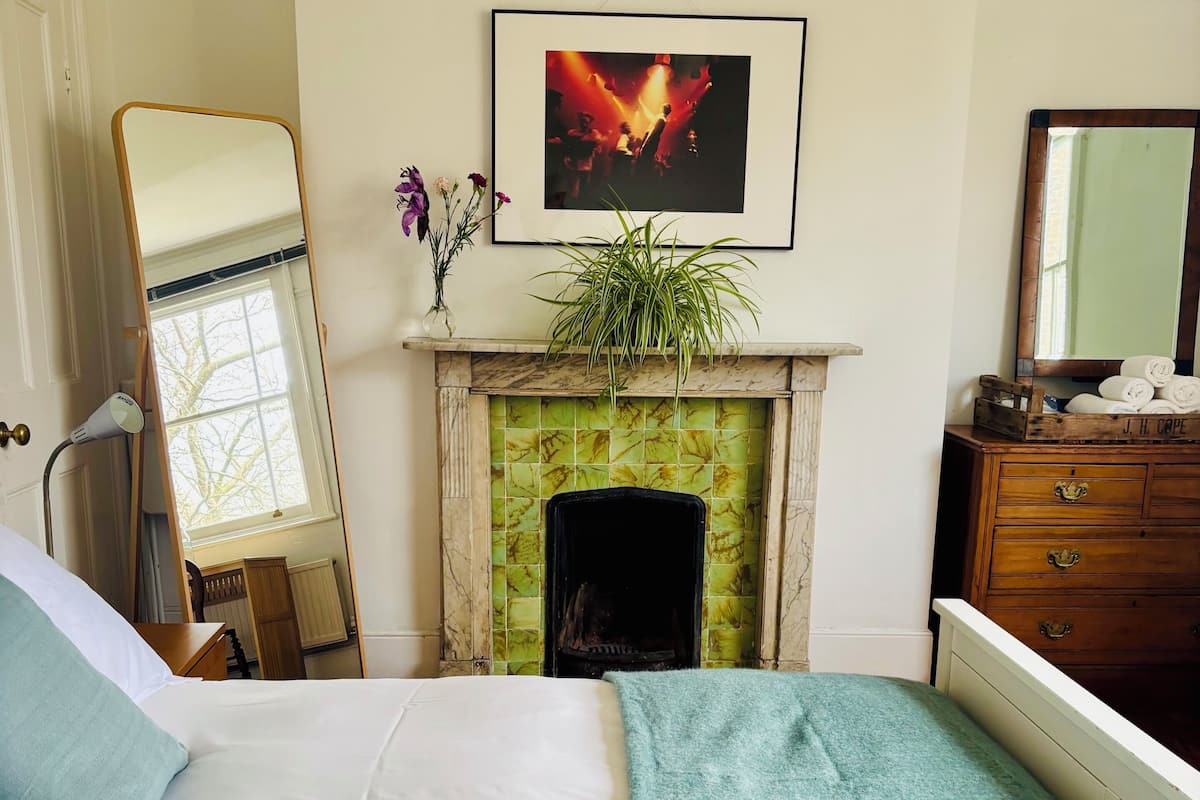 A bedroom with a bed, a mirror, a fireplace, and a framed picture on the wall.
