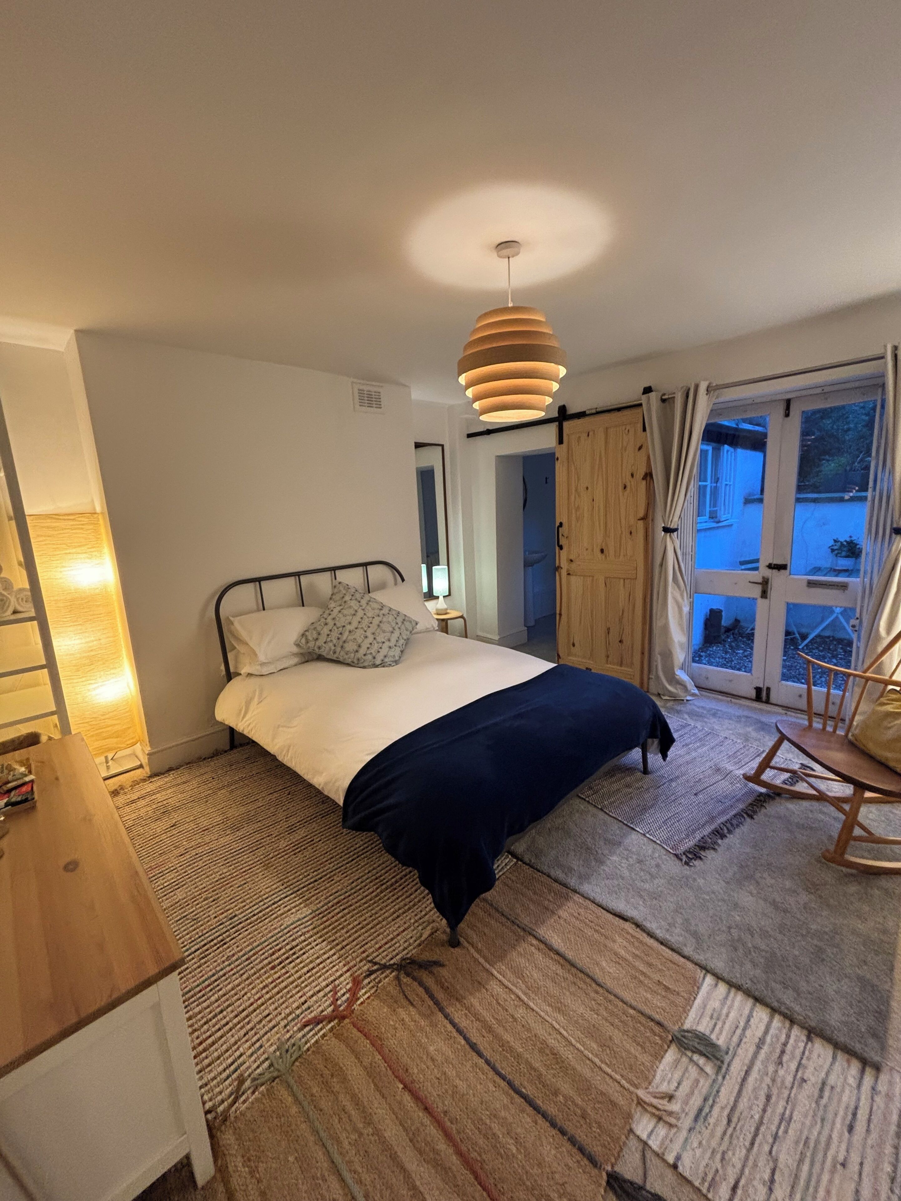 A bedroom with a bed, a wooden dresser, a hanging light fixture, and a view through a window.