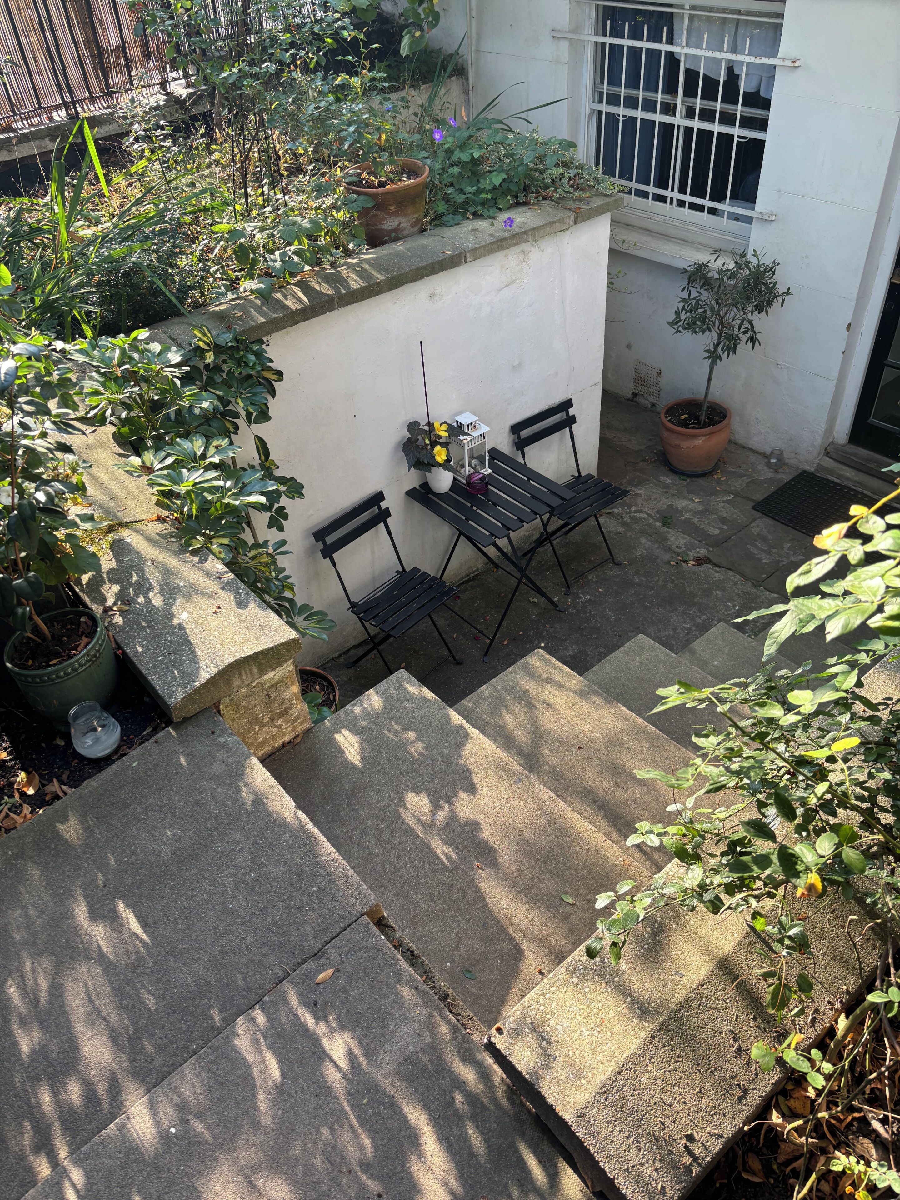 Un pequeño espacio al aire libre con una mesa y sillas, rodeado de plantas en macetas y una pared blanca.