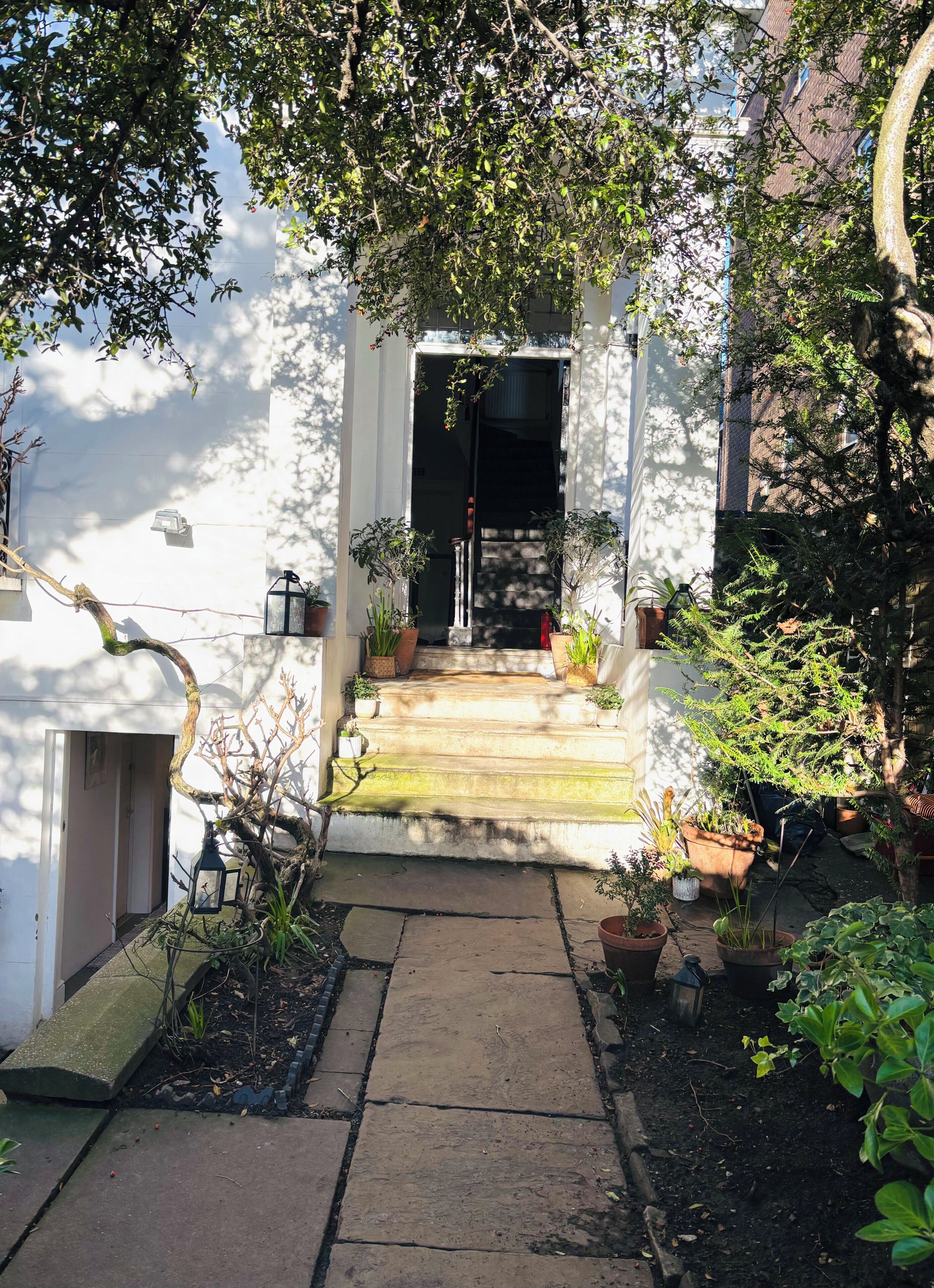 A stone pathway leading to a white building with a dark entrance, surrounded by potted plants and trees.