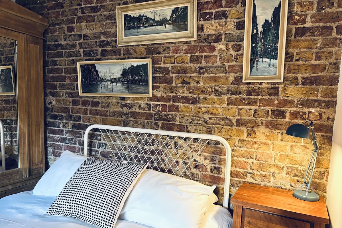 A room with a brick wall, a metal bed frame, white bedding, and a wooden nightstand with a lamp.