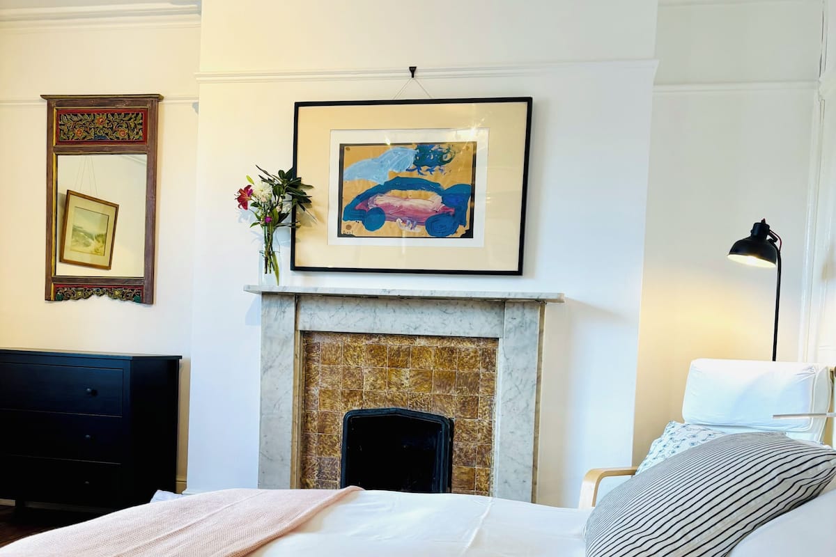 A hotel room with a large bed, a fireplace, a framed artwork above the mantel, a desk with a chair, and a lamp.