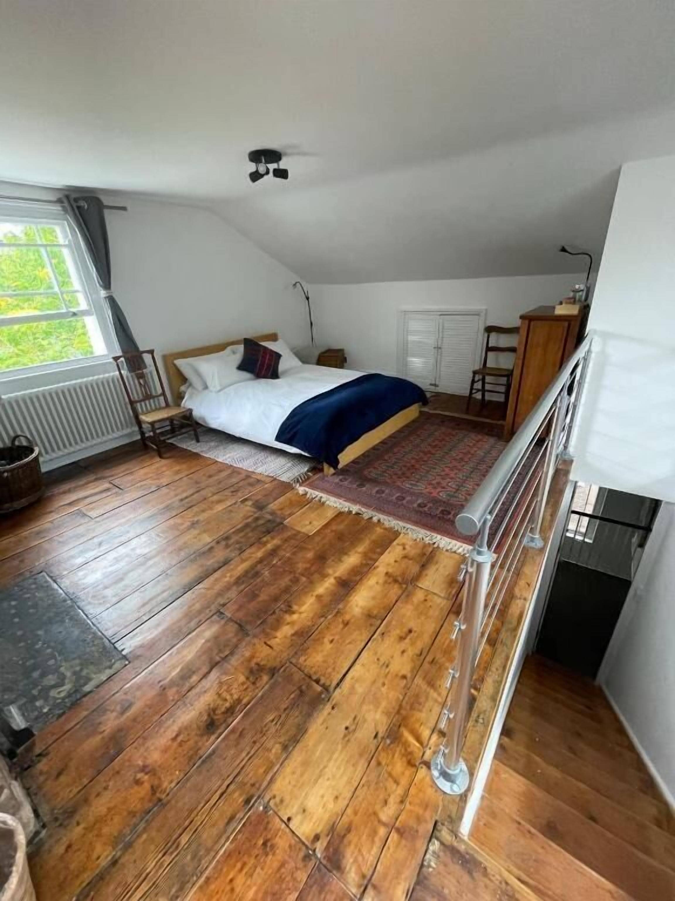 A bedroom with a wooden floor, a bed with a blue blanket, a window with curtains, and a ceiling fan.