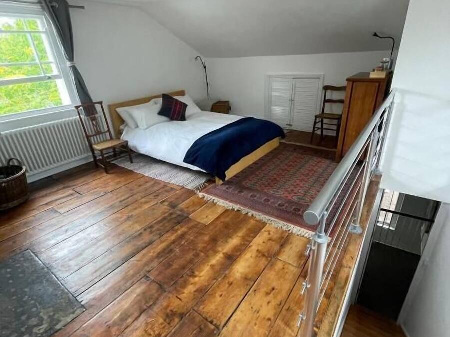 A bedroom with a wooden floor, a bed with a blue blanket, a window with curtains, and a ceiling fan.