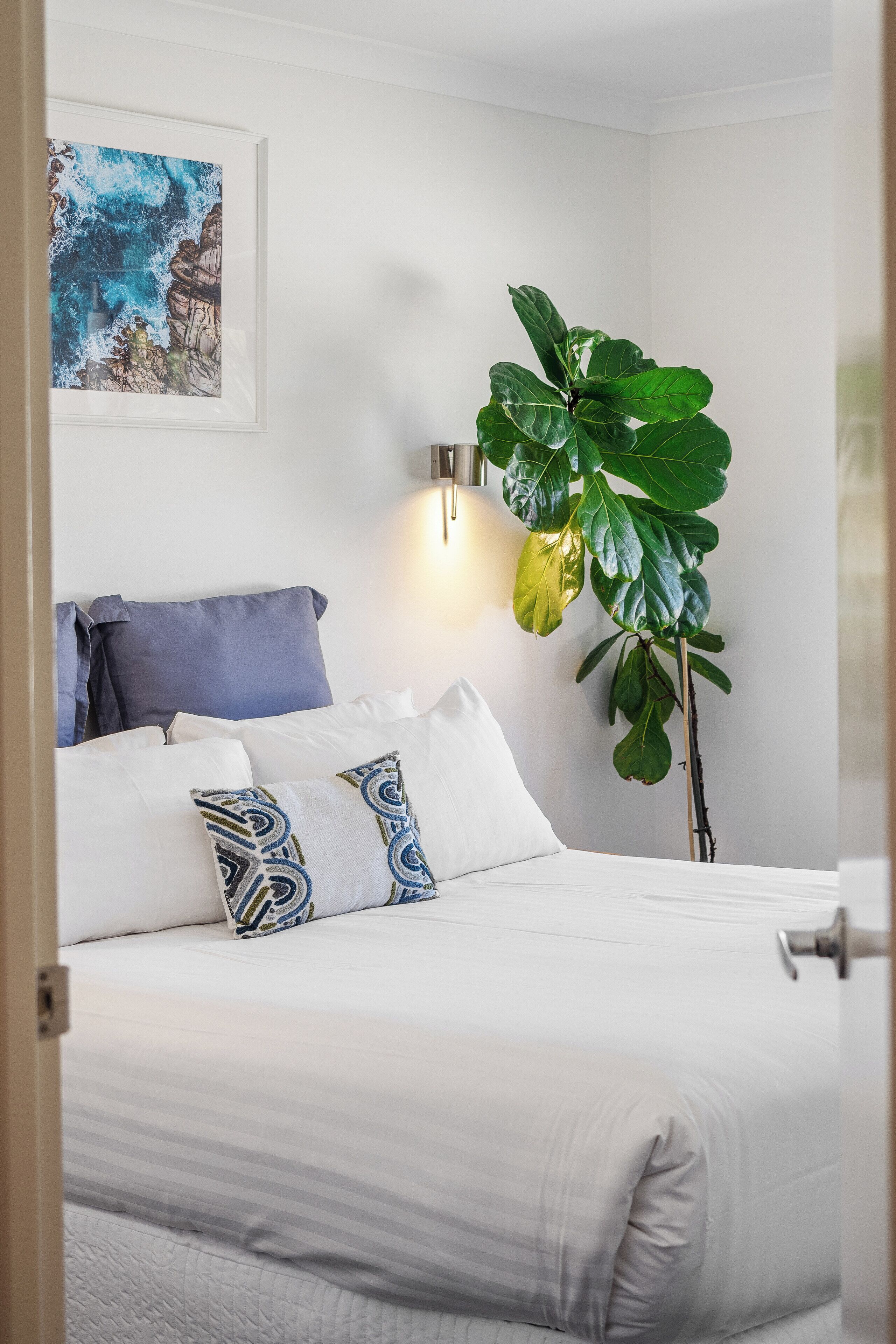 Una cama bien tendida con sábanas blancas y almohadas estampadas, una planta verde grande a un lado y una obra de arte enmarcada en la pared.