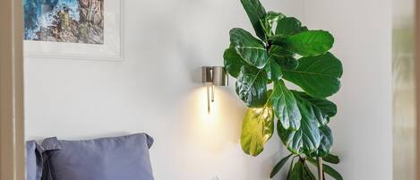 Una cama bien tendida con sábanas blancas y almohadas estampadas, una planta verde grande a un lado y una obra de arte enmarcada en la pared.