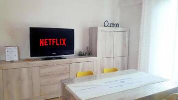 Ein Zimmer mit einem Fernseher, der 'NETFLIX' anzeigt, einem Holzschrank mit dem Schriftzug 'Cuina' auf der Oberseite und einem Tisch mit weißem Tischtuch.
