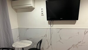 Una habitación pequeña con una televisión montada en la pared, una mesa con dos sillas y un equipo de aire acondicionado sobre la mesa.