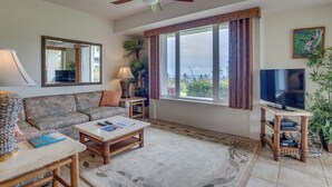 Ein Wohnzimmer mit einer Couch, Couchtisch, TV-Stand, Deckenventilator, großen Fenstern und Blick ins Freie.