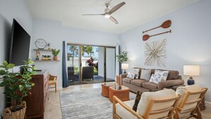 A living room with a sofa, chairs, a TV, a ceiling fan, and a view through a glass door to the outdoors.