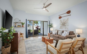 A living room with a sofa, chairs, a TV, a ceiling fan, and a view through a glass door to the outdoors.