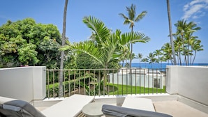 A balcony with white cushioned chairs and a table, overlooking a well-maintained garden with palm trees and a view of the ocean. - Royal Sea Cliff Kona 202 Ocean View 1 Bedroom (Kailua Kona)