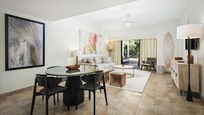 A modern living room with a round glass table, black chairs, a sofa, and a coffee table. There is a large abstract painting on the wall, a ceiling fan, and a view of the outdoors through a sliding door. - Royal Sea Cliff Kona 202 Ocean View 1 Bedroom (Kailua Kona)
