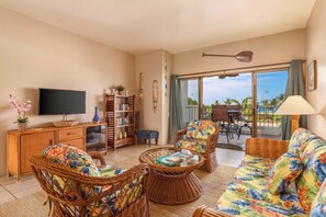 A living room with wicker furniture, a flat-screen TV, a bookshelf, and a view of the outdoors. - Royal Sea Cliff Kona 602 Ocean View 1 Bedroom (Kailua Kona)