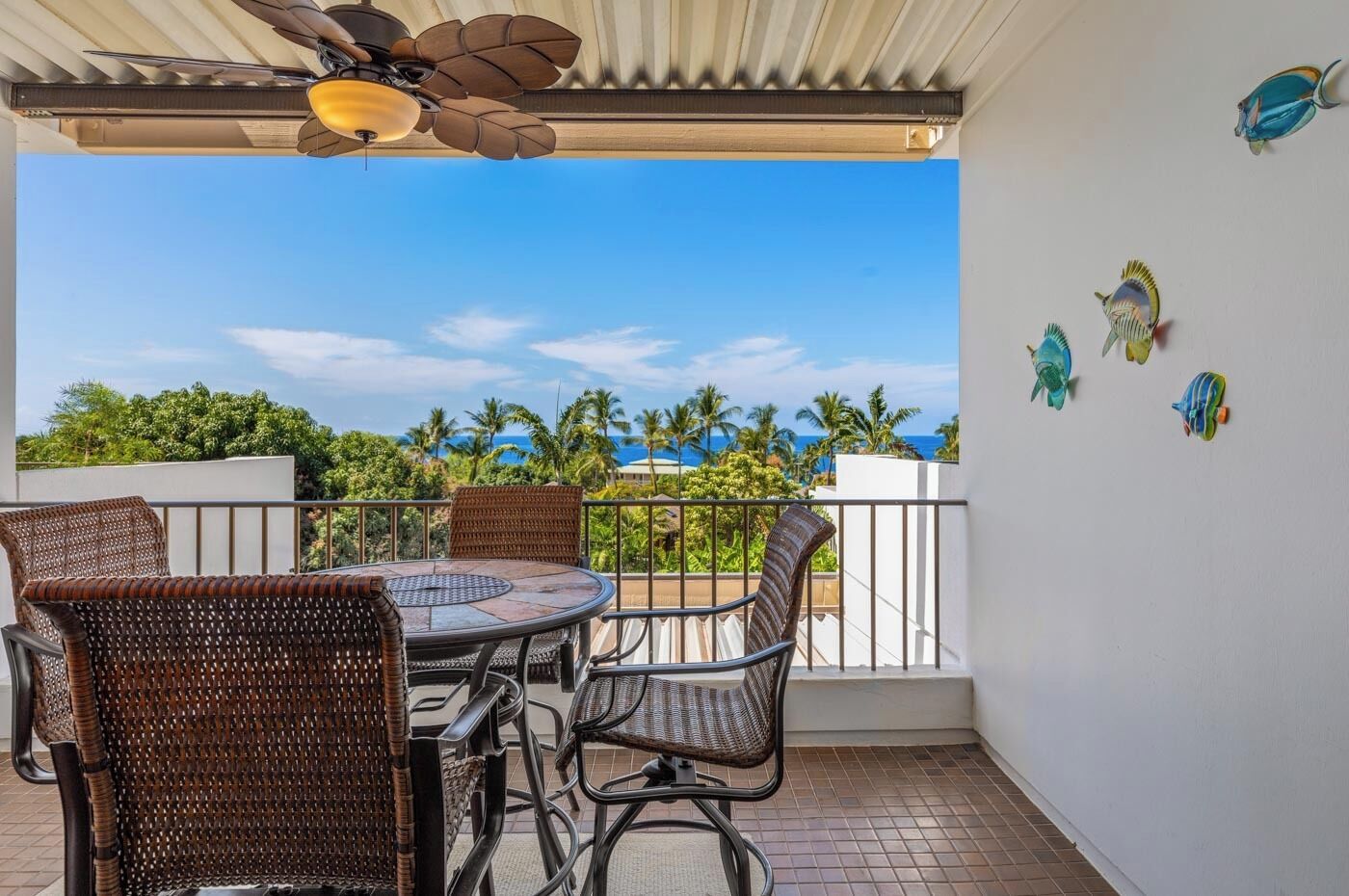 A covered outdoor seating area with a table and chairs, a ceiling fan, and a view of the ocean and palm trees.