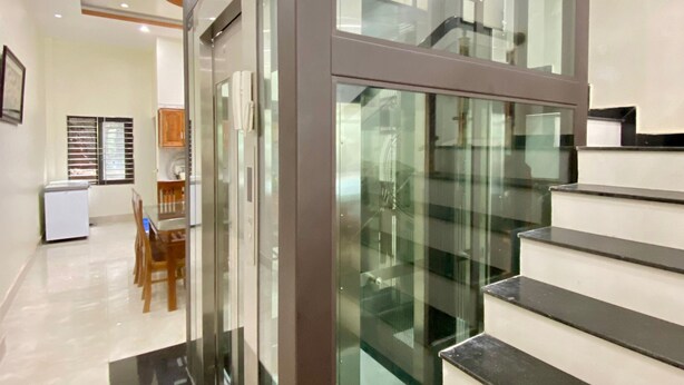 Un interno moderno con una scala in vetro, una zona pranzo con sedie e tavolo, e un angolo cottura con armadi.