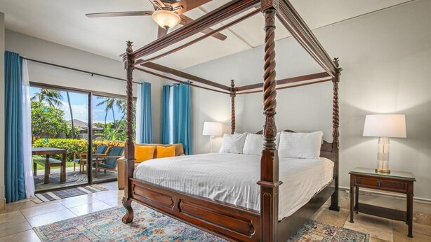 A four-poster bed with a wooden frame, a glass side table with a lamp, and a view of the outdoors through a sliding glass door.