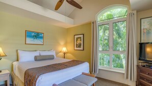 Une chambre d’hôtel avec un grand lit, un ventilateur de plafond, une télévision, une fenêtre avec des rideaux et un bureau avec une lampe.