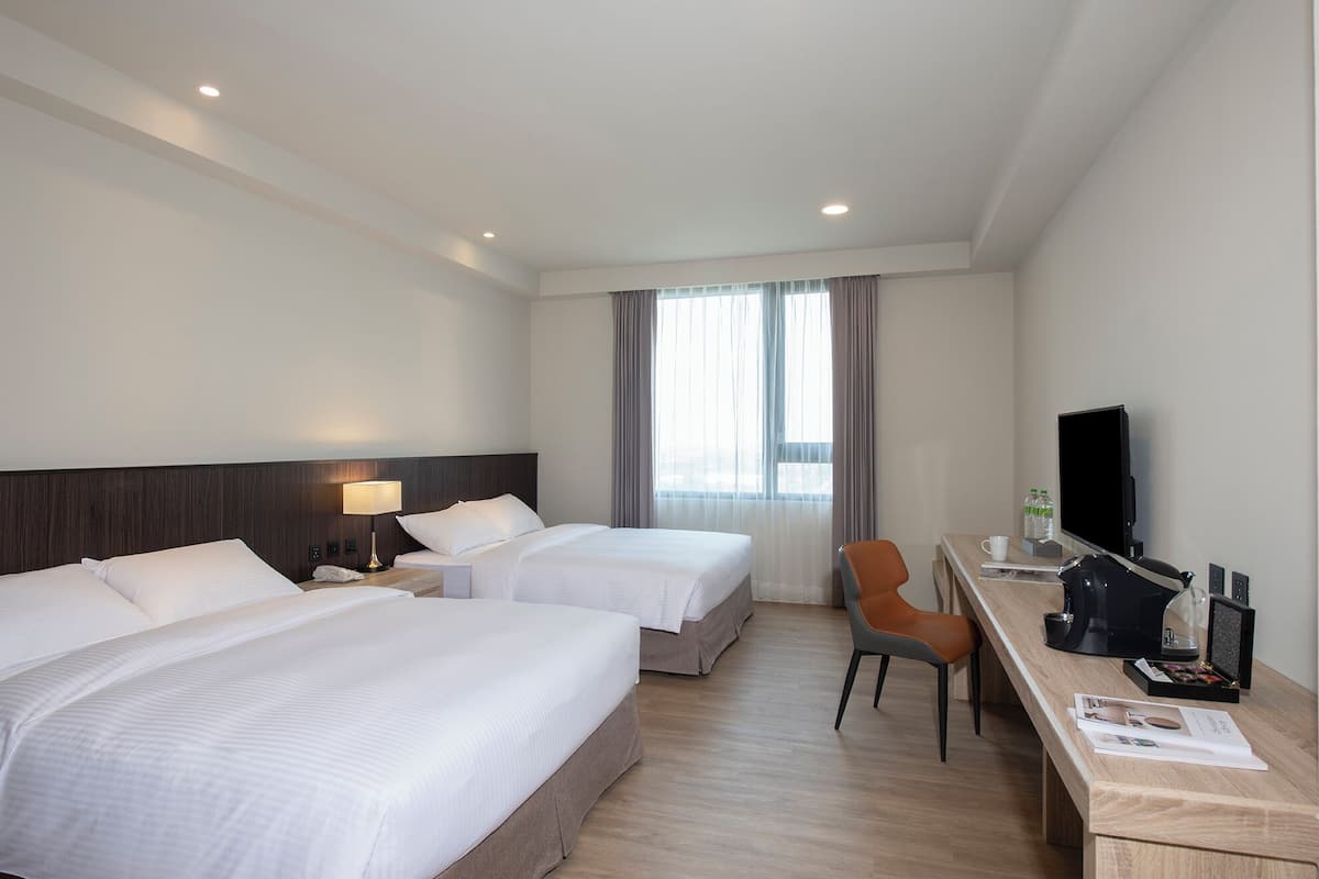 Quarto de hotel com duas camas, uma mesa com cadeira, televisão e janela com cortinas.