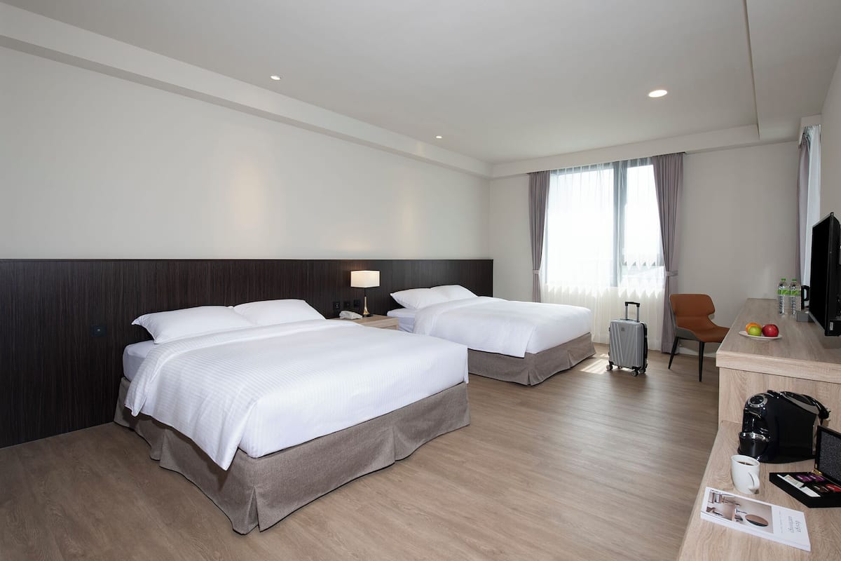 Quarto de hotel com duas camas, uma escrivaninha com cadeira, televisão e uma janela com cortinas.