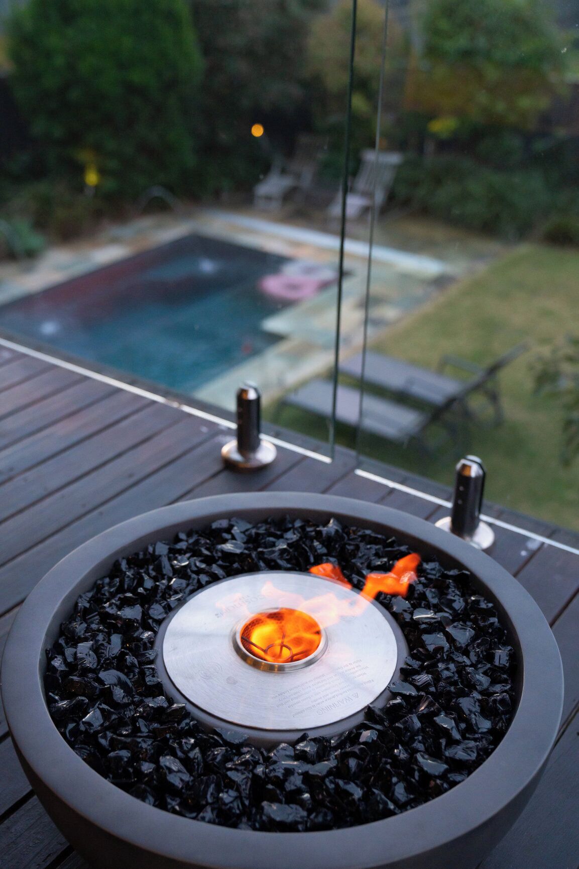A circular outdoor fire feature with a central flame, surrounded by black stones, on a wooden deck.