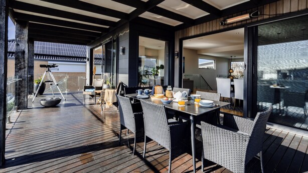 Un área de comedor al aire libre moderna con terraza de madera, sillas de estructura metálica y cocina con paredes de vidrio al fondo.