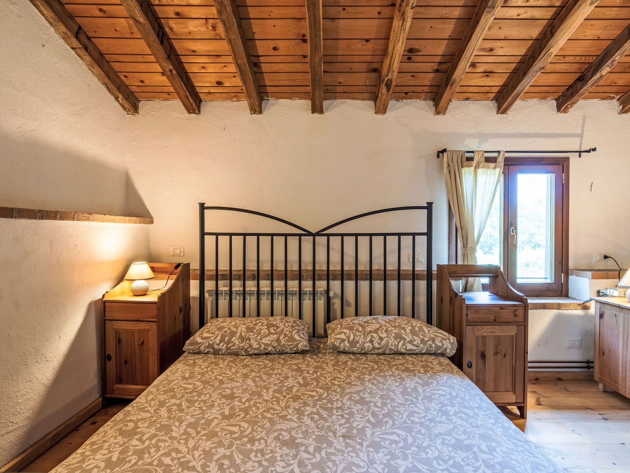A bedroom with a wooden ceiling, a bed with patterned bedding, a nightstand with a lamp, and a window with curtains.
