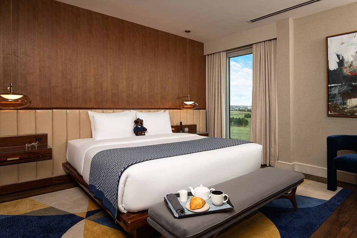 A hotel room with a large bed, a bedside table with a lamp, a view of the outdoors, and a breakfast tray on the bed.
