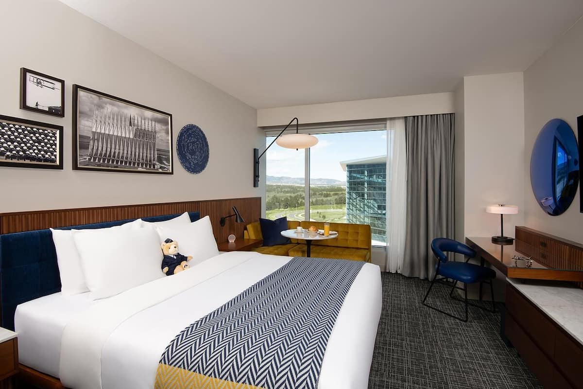 Habitación de hotel con una cama grande, un escritorio, una silla, una mesita y un ventanal con vistas a un edificio y un paisaje.