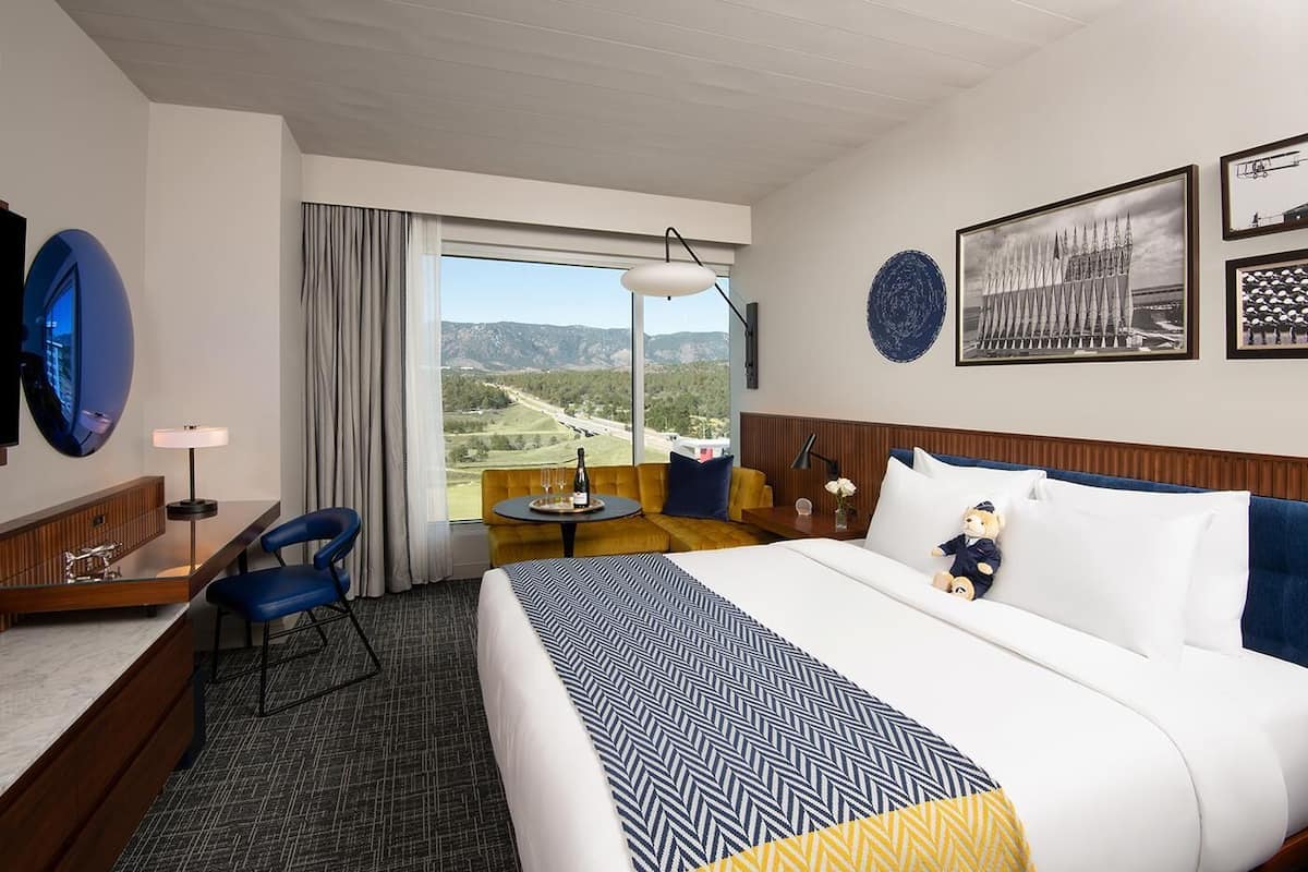 Habitación de hotel con una cama grande, un escritorio con una silla, una mesita con una botella y vasos, un sofá, y vistas a un paisaje a través de un ventanal amplio.