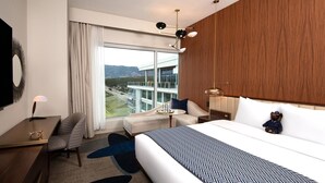 A hotel room with a large bed, a desk, a chair, a sofa, and a view of a building and mountains.
