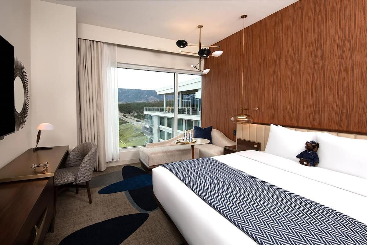 Habitación de hotel con una cama grande, un escritorio, una silla, un sofá y vistas a un edificio y montañas.