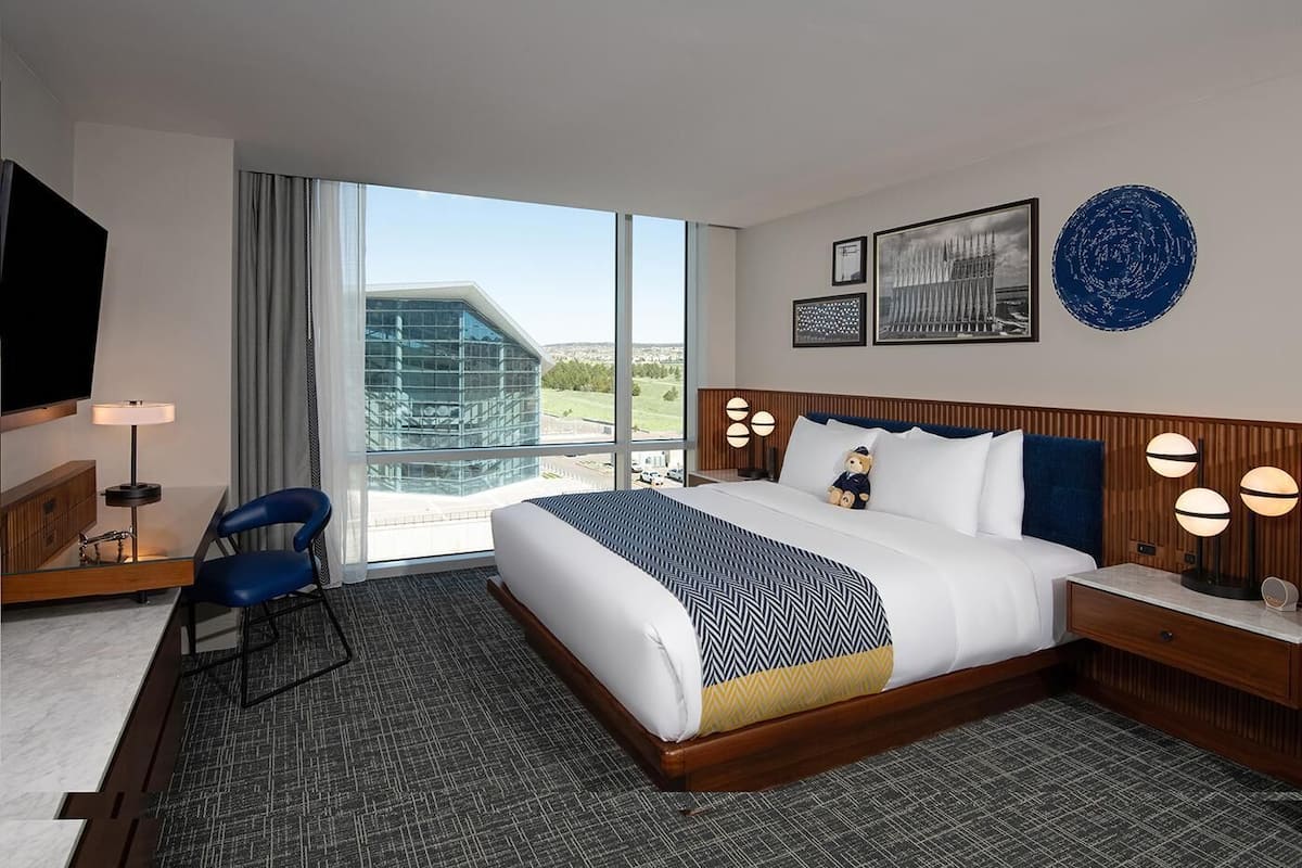 Habitación de hotel con una cama grande, un escritorio con una silla, televisión y vista a un edificio desde la ventana.