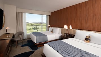 A hotel room with two beds, a desk, a chair, and a large window with a view of the outdoors.