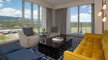 A modern living room with a sofa, armchair, and coffee table, large windows with a view of mountains and a road, and a balcony with a railing.