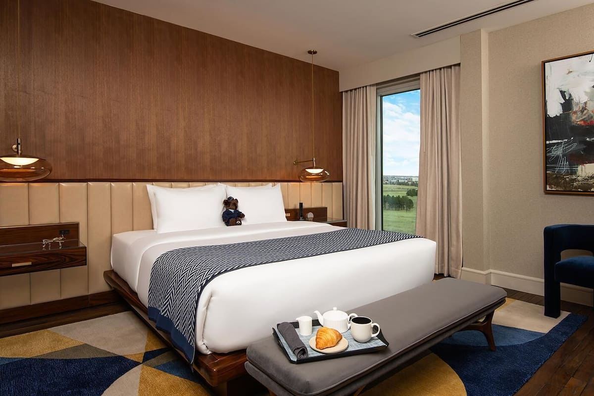 Habitación de hotel con una cama grande, una mesita de noche con lámpara, vista al exterior y una bandeja de desayuno en la cama.