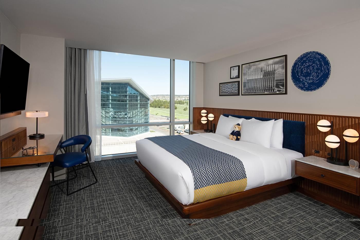 A hotel room with a large bed, a desk with a chair, a TV, and a view of a building through the window.