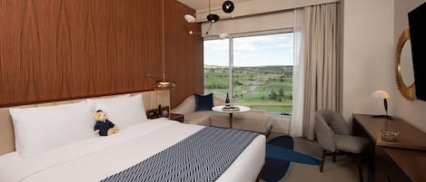 A hotel room with a large bed, a sofa, a desk, and a chair. There is a window with a view of the countryside.