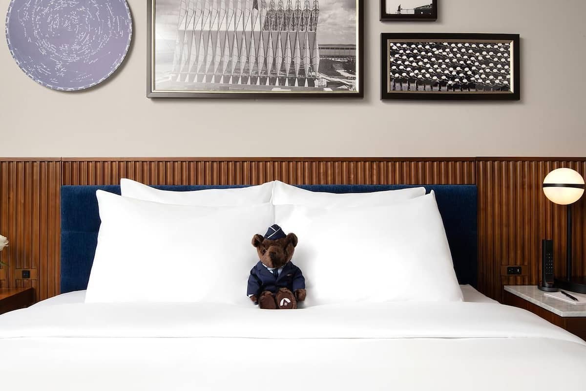 Una habitación de hotel con una cama grande, un osito de peluche vestido de piloto y cuadros en la pared.
