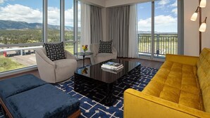 A modern living room with a sofa, armchair, and coffee table, large windows with a view of mountains and a road, and a balcony with a railing.