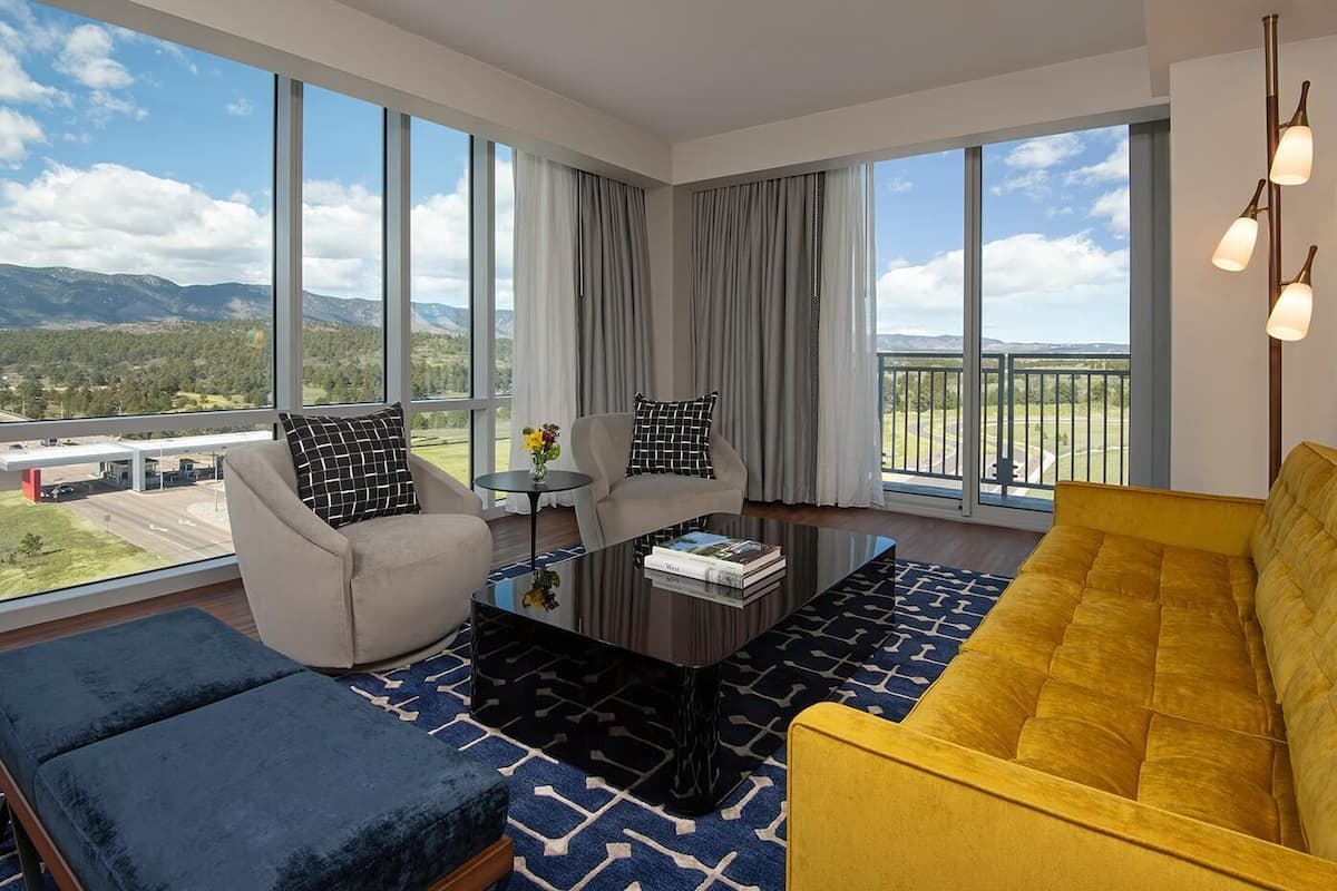 Una sala de estar moderna con sofá, sillón y mesa de centro, ventanas amplias con vistas a montañas y una carretera, y un balcón con barandilla.