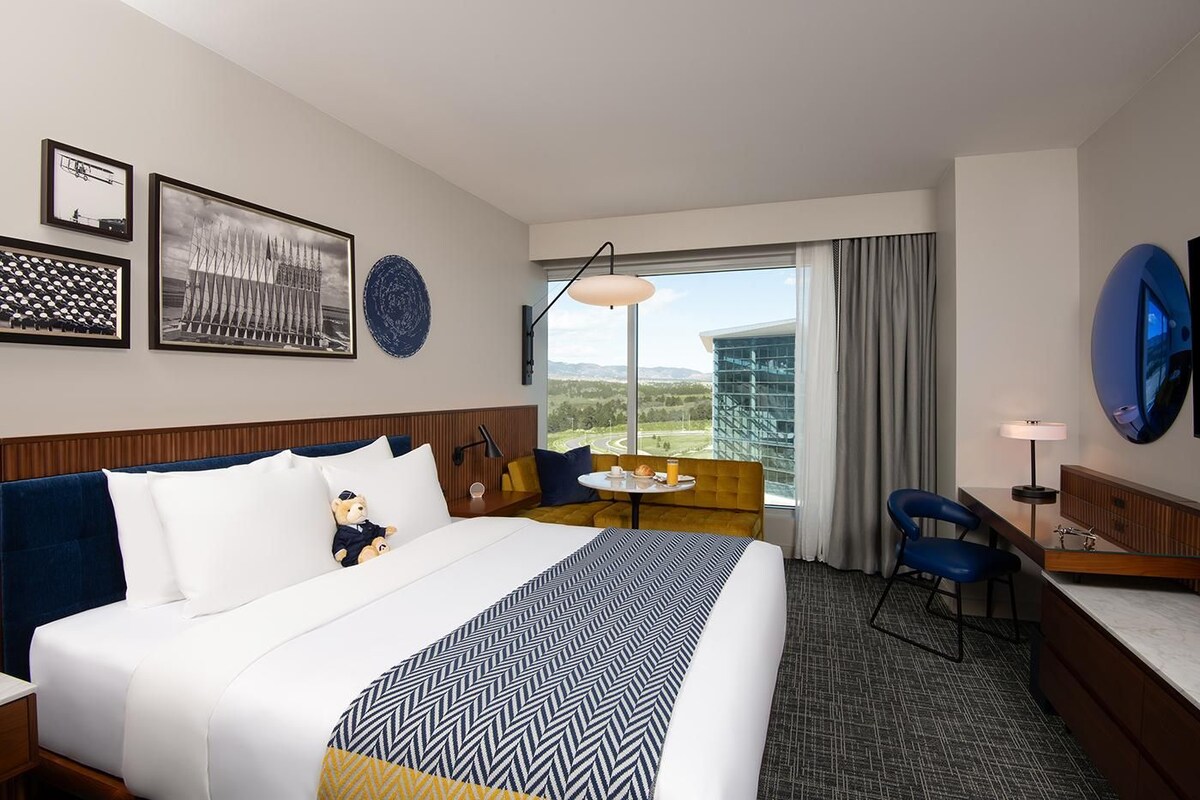 A hotel room with a large bed, a desk, a chair, a small table, and a large window with a view of a building and landscape.