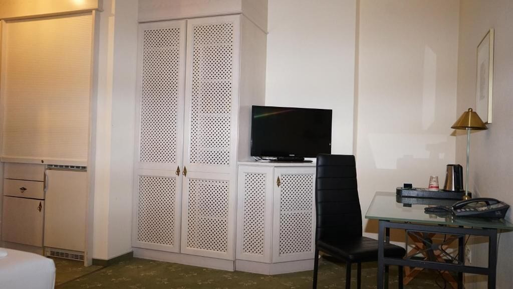 A hotel room with a TV on a stand, a desk with a chair, and a wardrobe with a perforated design.