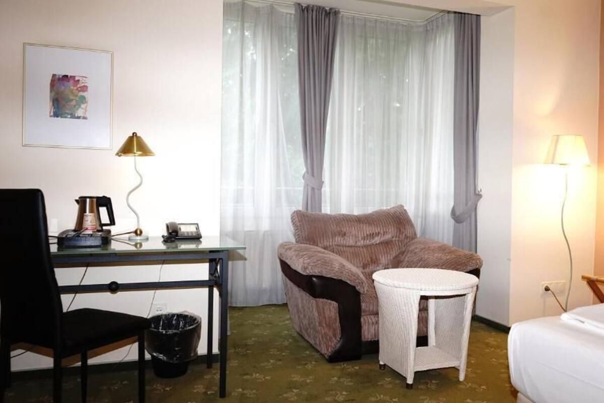 A hotel room with a bed, a desk with a chair, a small round table, and a window with curtains.