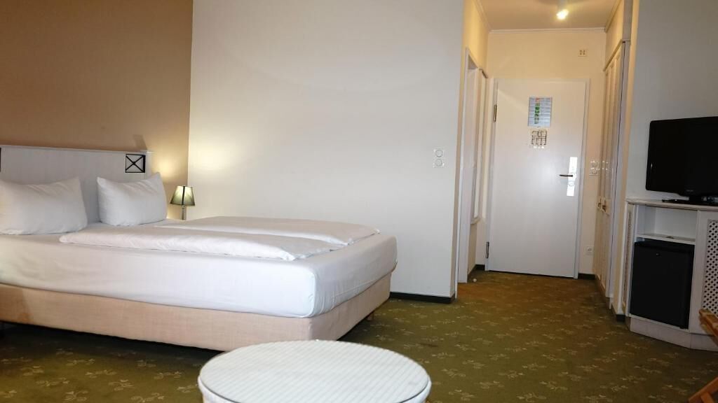 Habitación de hotel con una cama grande, una mesita de noche, un televisor y una puerta que da a un pasillo.