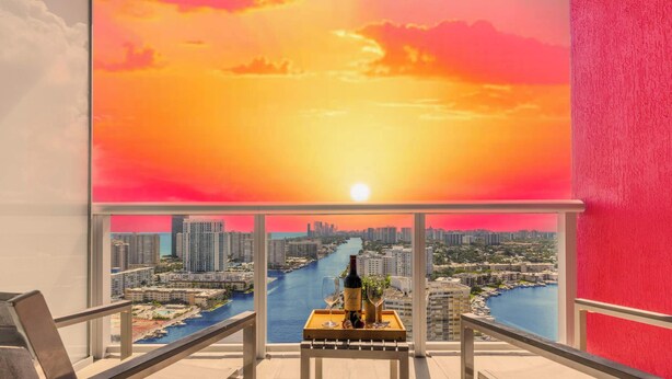 A balcony with a city view at sunset, a table with a bottle and glasses, and a chair. - Beautiful Studio, Amazing View Balcony (Hallandale Beach)