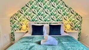 Un lit bien fait, avec une couverture verte et deux oreillers, une tête de lit blanche et un mur orné d’un motif végétal.