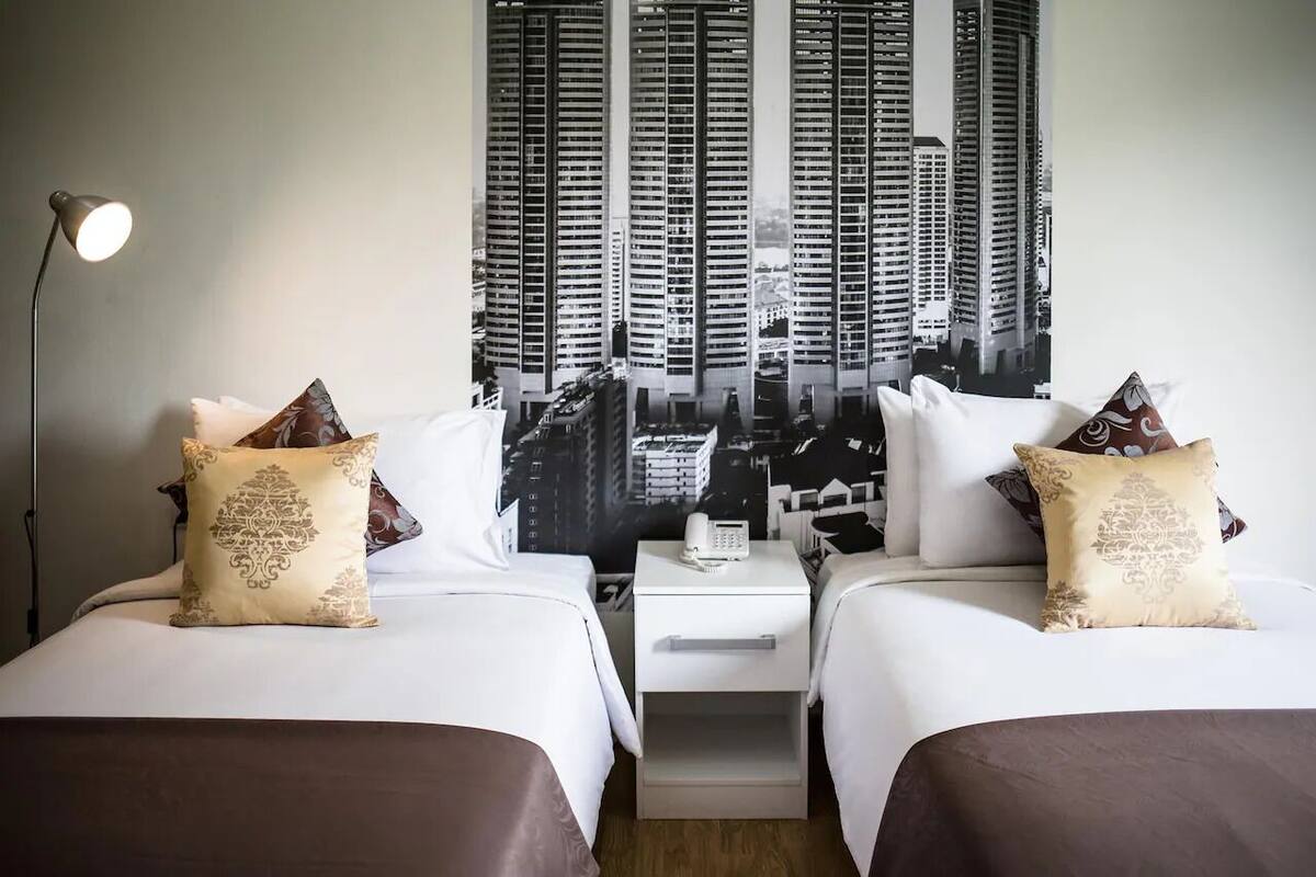Habitación de hotel con dos camas, una mesita de noche, una lámpara y un mural con vistas a la ciudad.